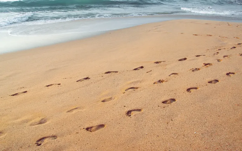 Symbolbild: Fußspuren im Sand eines Strandes, dahinter flache Wellen