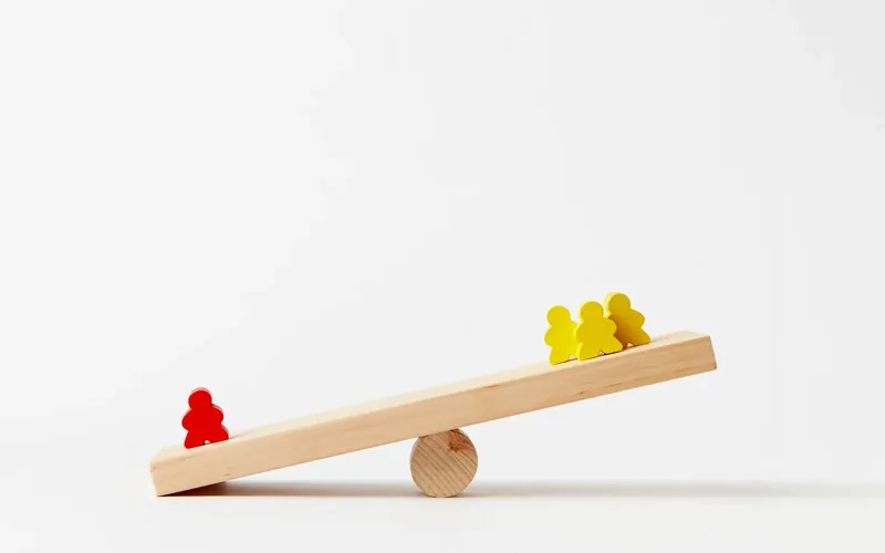 Symbolbild: Holzwippe mit einer roten Spielfigur auf der einen und drei gelben auf der anderen Seite
